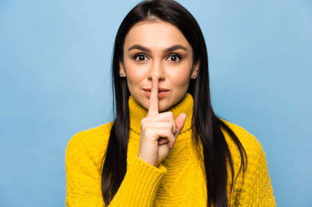 Portrait of cute caucasian young woman, shows gesture shhh, do not tell secret information, stands...
