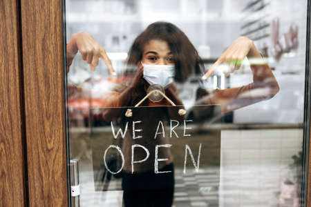 Curly female African American small business owner in medical mask points at a signboard WE ARE OPEN...