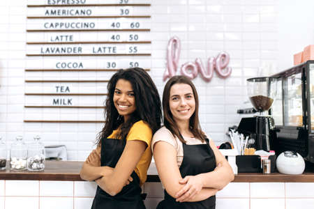 Portrait of two female waitress of a coffee shop, African American and Caucasian, stands near the bar counter backs to each other with arms crossed, looking and smiling at the cameraの写真素材