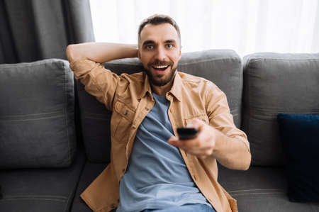 Excited guy watch on tv sport channel, sitting and relaxed on the couch holds a tv remote control, surprised with results of match, leisure time conceptの写真素材