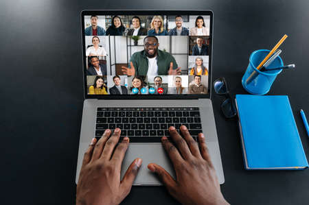 Online business meeting. Multiracial business people working from home by a video conference. Top view at a laptop screen with webcam shots of diverse people and hands of a black man on a keyboardの写真素材