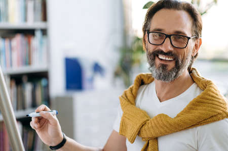 Portrait of a happy male employee or teacher in eyeglases, holds a marker in hand, writing on flipchart financial graphs or theme of lecture, looks and smiles at cameraの写真素材