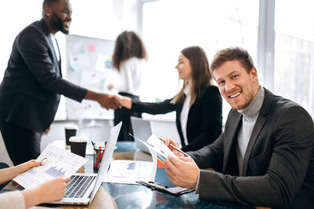 Portrait of smiling male manager in formal wear at briefing meeting. Successful business man looks directly at the camera and smile, in the background, colleagues shaking handsの写真素材