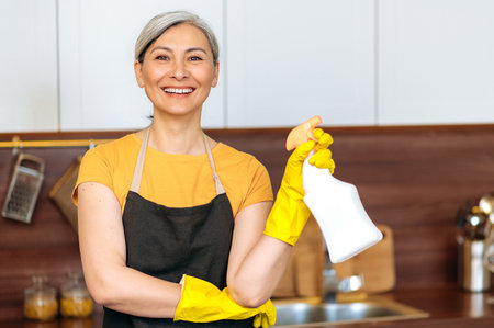 Satisfied smiling mature asian housewife or cleaning lady in yellow gloves and an apron, holding...