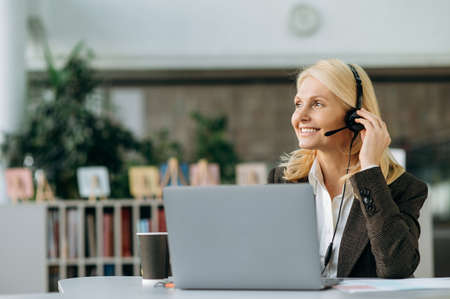 Elegant happy business woman is working as operator of call center, sitting at the desk in office. Mature female employee in headset and stylish suit consulting clients, smilingの写真素材