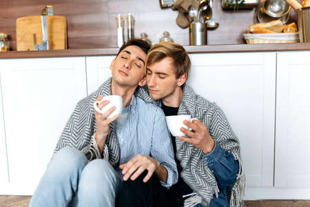 Homosexual relationship. Two boyfriends guys in love spend time together, they hugging tenderly each other, covering with plaid while sitting on the floor at kitchen, with cups of coffeeの写真素材