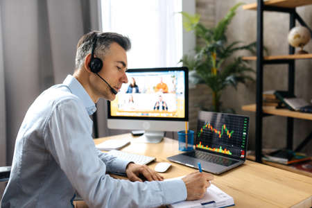 Online lesson or business webinar. Successful man sits at the work desk makes notates during online meeting, on a computer screen are different multiracial people gathered in an online lecture byの写真素材