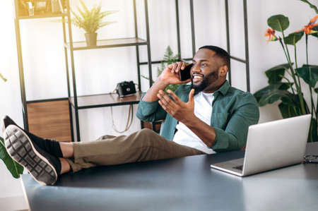 Happy African American young man, freelancer or modern ceo, is relaxing at workplace with legs thrown on table, has a pleasant phone conversation with a friend or girlfriendの写真素材