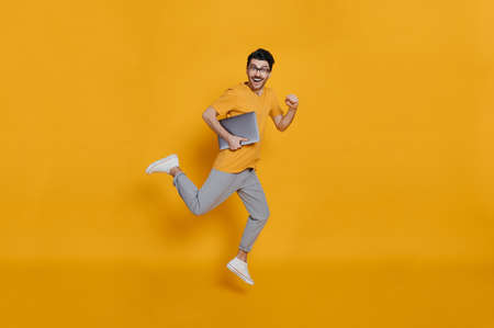 Full length photo of a cheerful funny caucasian smart young man in eyeglasses, stylishly dressed, holding laptop, smiling and jumping high running on isolated orange backgroundの写真素材