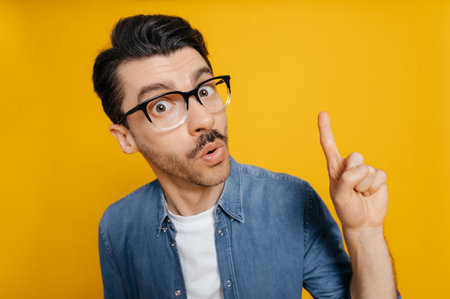 Distorted portrait of amazed surprised caucasian guy in a glasses and in an orange t-shirt, shocked looking at the camera, shows IDEA gesture, standing against an isolated orange backgroundの写真素材