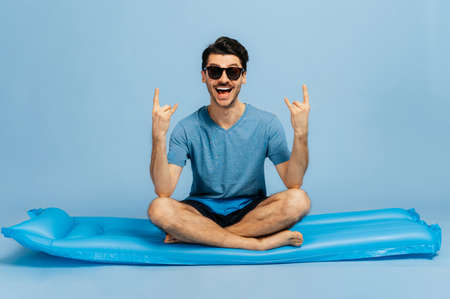Cheerful caucasian good looking happy guy in sunglasses sitting on blue inflatable mattress in lotus...