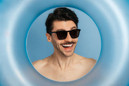 Close-up photo of a shirtless cheerful happy caucasian young man in a sunglasses looks through...