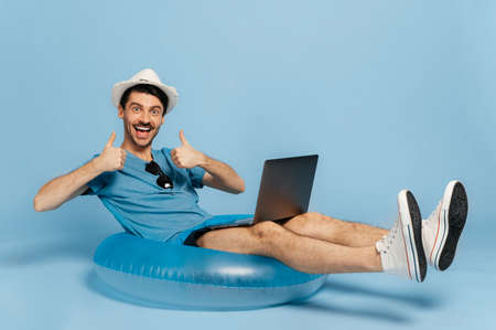 Happy excited smiling freelancer guy in blue t-shirt and hat, sitting on an inflatable blue ring, working remotely using a laptop, looks at the camera, gesturing thumbs up, blue isolated backgroundの写真素材