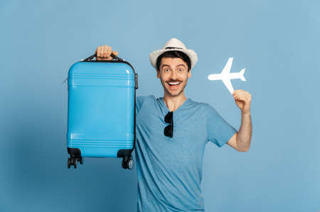 Happy satisfied caucasian guy in a hat holds a blue suitcase and a small airplane, looking directly at the camera, smiling happily, in anticipation of a long-awaited vacation, isolated blue backgroundの写真素材