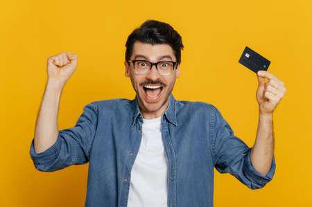 Young excited positive caucasian man holds credit card, rejoices in success, victory, profit,...