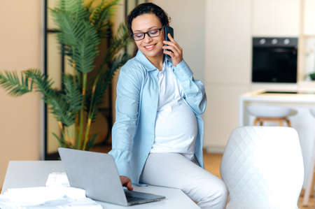 Busy good looking confident pregnant mixed race adult woman is sitting on her work desk, having pleasant cellphone conversation uses and looking at the laptop laptop, smilingの写真素材