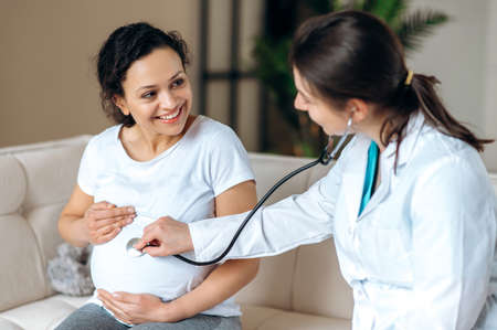 Health of a pregnant woman and a baby. Female doctor conducts checkup of happy joyful smiling pregnant mixed race woman, listening to babys heartbeat using stethoscope, sitting on sofa in living roomの写真素材