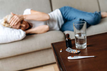 Defocused sick adult woman at home on the couch. A middle-aged caucasian ill woman have headache, clutching her head, a glass of water on the table and a thermometer with pills, need a treatmentの写真素材