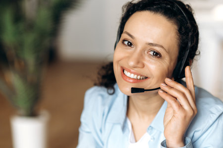 Close-up of mixed race attractive confident adult woman in headset, sitting in office, working as operator of call center or support service, looking directly at the camera and smiling friendlyの写真素材