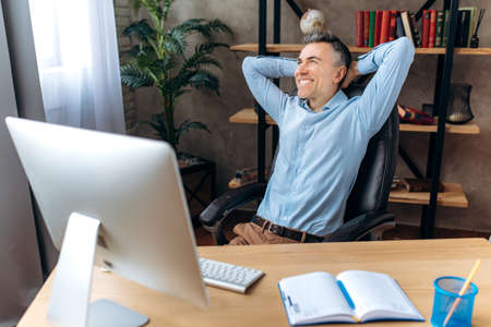 Happy satisfied businessman in stylish formal clothes, sitting at his workplace in the office, hands...