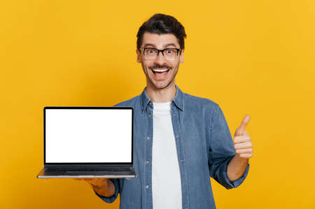Amazed excited cheerful caucasian unshaven guy in glasses and denim shirt holds open laptop with blank white screen, showing thumbs up gesture, standing on isolated orange background, friendly smilingの写真素材