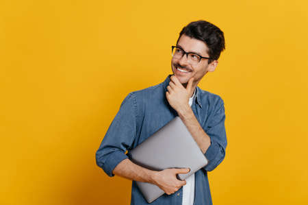 Attractive confident smart guy in glasses and in a denim shirt, holding a laptop at hand, looks thoughtfully towards empty space aside touching his chin, smiling, isolated orange background,copy spaceの写真素材