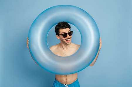 Excited cheerful happy caucasian young man without t-shirt and in a sunglasses looks through inflatable ring at the camera, stands over blue isolated background and joyfully smiling, summer vacationの写真素材