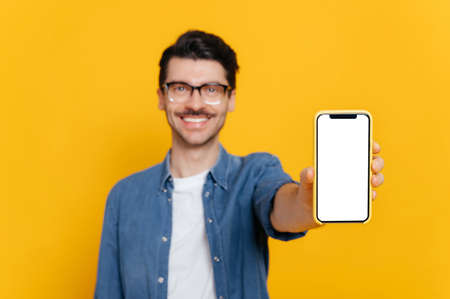 Defocused caucasian unshaven smiling guy with glasses in denim shirt, looks at the camera, holds smartphone with blank white screen in hand, copy space, isolated orange backgroundの写真素材