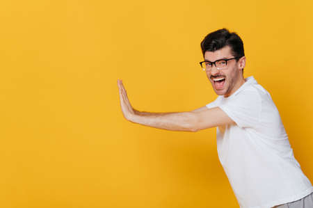 Screaming amazed excited caucasian guy wearing glasses and white basic t-shirt gesturing hands towards empty space looks at camera while standing against isolated orange backgroundの写真素材