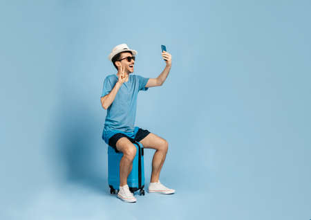 Cheerful guy dressed in summer clothes, wearing sunglasses and a hat, sits on a blue suitcase, takes a selfie on the phone or communicates on a video call, goes on vacation, blue isolated backgroundの写真素材
