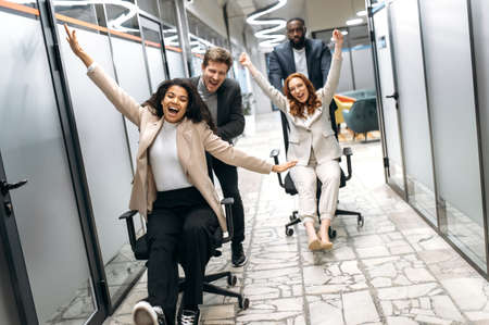 Joyful male and female colleagues take a break from work, having fun. Happy multiracial office employees in formal wear making a race on chairs, smiling and laughingの写真素材
