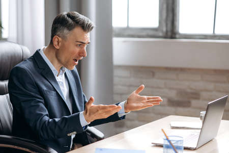 Shocked annoyed caucasian busy business man in suit, looking at a laptop in confusion, received unexpected news, failed a deal, shouts, gesturing with hands, sitting at the work desk at the officeの写真素材