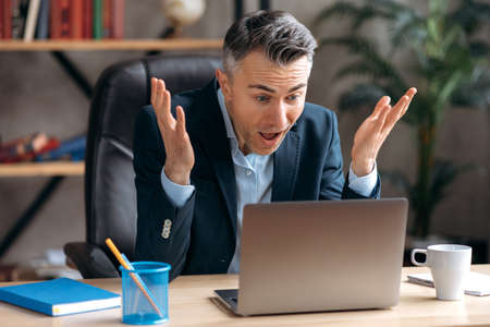 Surprised caucasian successful senior grey haired man, manager or lawyer, being in the office, delighted, happy with success, good profit, looking at a laptop with an open mouth, gesturing with hisの写真素材