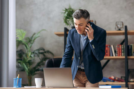 Busy successful handsome grey haired caucasian businessman, ceo, in formal suit, stands at his modern office, talking on a mobile phone with a colleague or clients, uses a laptop and browsing internetの写真素材