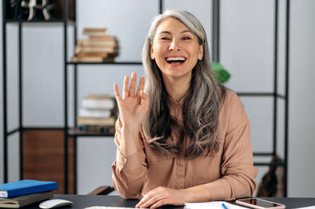 Joyful beautiful confident gray-haired Asian woman, freelancer, business lady, manager, chatting on a video call, conducts online briefing with colleagues or friends, gesturing with her hand, smilingの写真素材