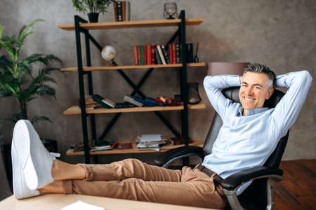 Successful handsome mature caucasian businessman or manager, freelance relaxes while sitting with his legs thrown on his desk, looking at the camera, with hands behind his head, smiling friendlyの写真素材