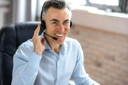 Portrait of successful, confident handsome senior businessman, manager, or call center worker, sits in a modern office, uses headphones for communicate with client, looks at the camera, friendly smileの写真素材