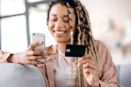 Defocused african american joyful smiling girl holding smartphone and bank card, shopping online,...