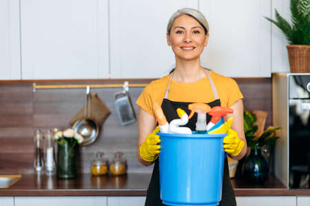 Friendly mature gray-haired Asian cleaning lady or housewife in gloves and an apron is standing in the kitchen, holding large bucket of detergents and rags, looking at the camera, smilingの写真素材