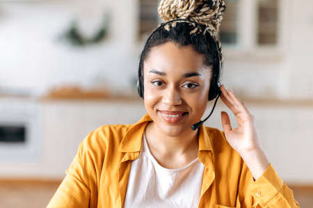 Close-up of African American an attractive confident young woman in headset, sitting in office, working as operator of call center or support service, looks directly at the camera and smiling friendlyの写真素材