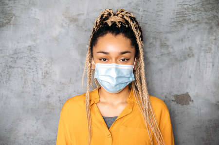 Precautions. African american pretty brunette woman with dreadlocks, in casual wear standing indoors on gray background with protective medical mask on her face and looking directly into the cameraの写真素材