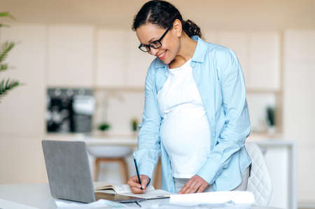 Beautiful successful pregnant mixed race woman wearing glasses, standing near workplace, having video call with colleague or watching video tutorial, receiving consultation, taking notes, smilingの写真素材