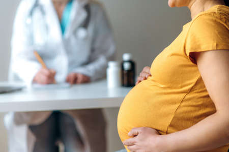 Pregnant woman in yellow t-shirt, hugging her belly, sits at the reception at the hospital, is undergoing treatment or get consultation, the doctor prescribes medications. Pregnancy healthcare conceptの写真素材