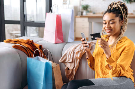 Beautiful satisfied african american girl with dreadlocks sits on sofa at home, using smartphone and...