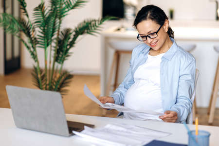 Pregnant mixed race female. Attractive joyful successful pregnant woman wearing glasses, creative designer or manager, working remotely, checking design project, studies documents, smilingの写真素材