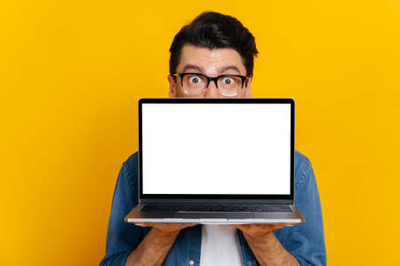 Amazed excited caucasian guy with glasses peeking out from behind laptop, looks surprised at camera, stands on isolated orange background, holds an open laptop with blank white screen, copy spaceの写真素材