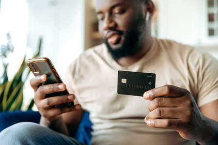 Defocused happy young African American bearded guy, in casual clothes, sitting on the couch at home,...