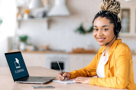 E-learning concept. Happy African American female student with dreadlocks wearing headset, sits at...