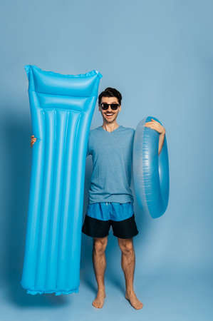 Full-length of amazed happy caucasian shirtless guy, standing with blue inflatable ring and mattress on an isolated blue background in sunglasses looks at camera and smiling, vacation conceptの写真素材