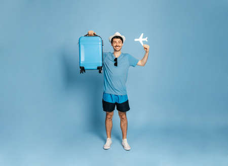 Full-length photo of a happy excited caucasian guy in a hat holds a blue suitcase and a small airplane, male tourist looks directly at the camera, smiling happily, stand on an isolated blue backgroundの写真素材